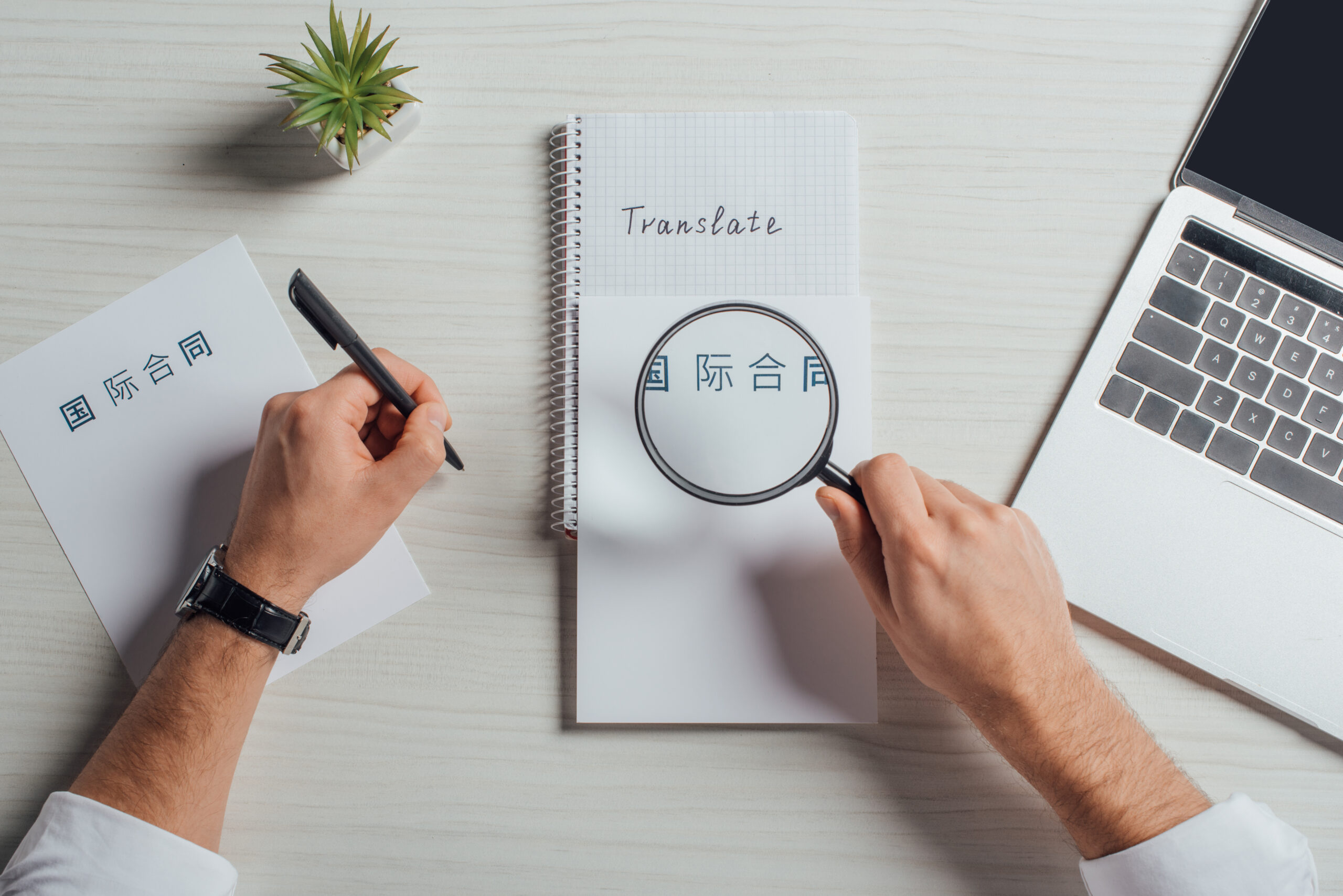 A person using a magnifying glass to examine Chinese characters on a notebook with the word 'Translate' on top, with a pen and laptop beside them.