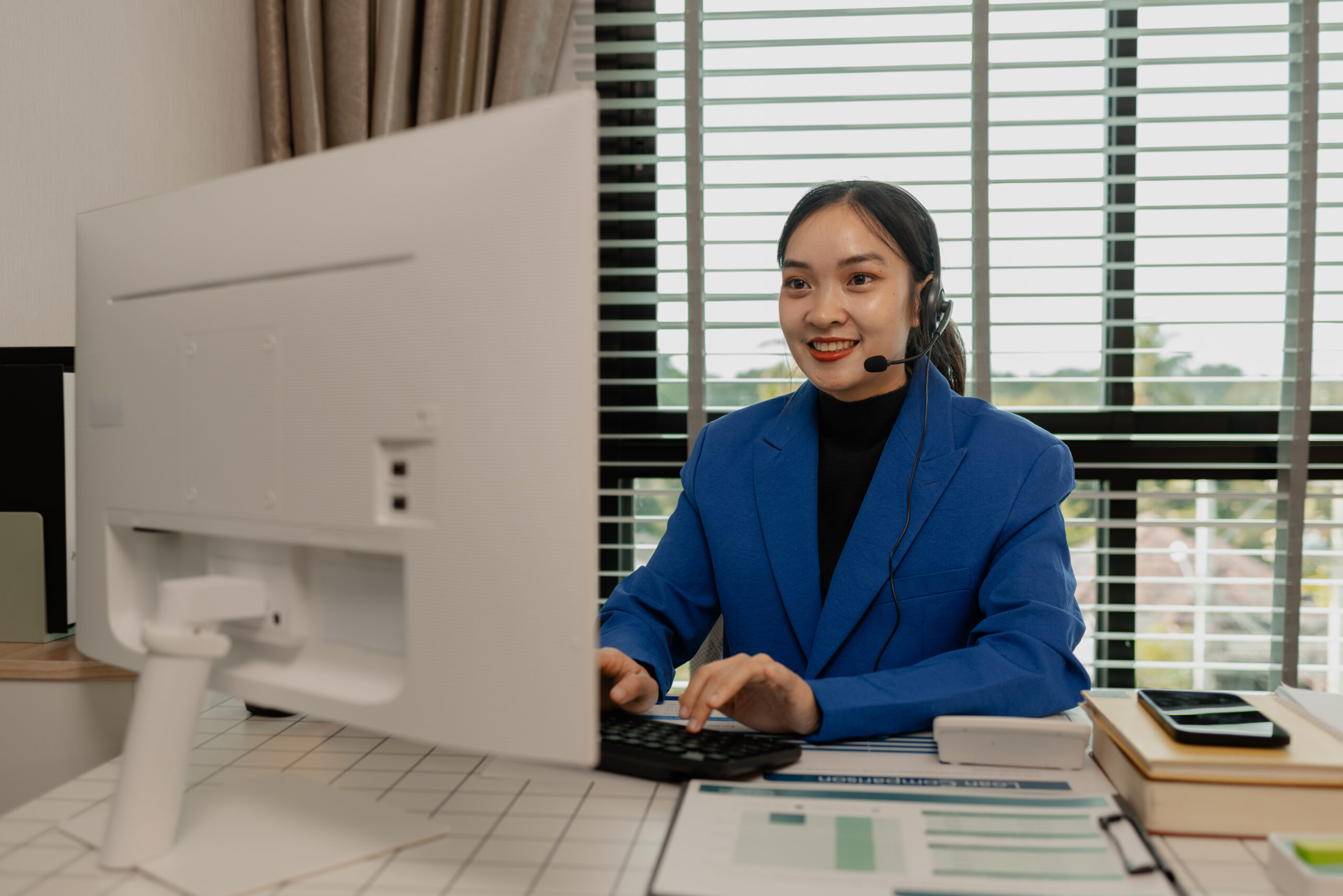 A woman in a blue blazer wearing a headset, smiling while typing on a keyboard in front of a desktop computer with a view of the outdoors.