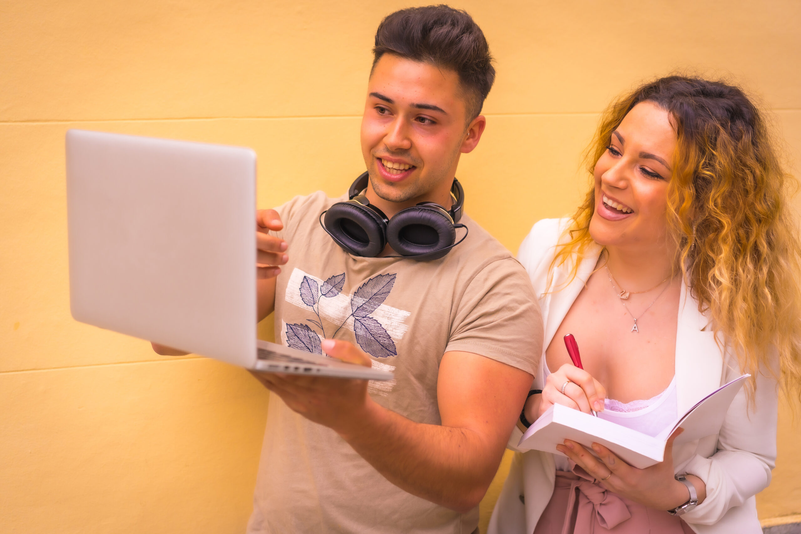 A man holding a laptop and showing something on the screen, while a woman smiles and writes in a notebook, both standing in front of a yellow wall.